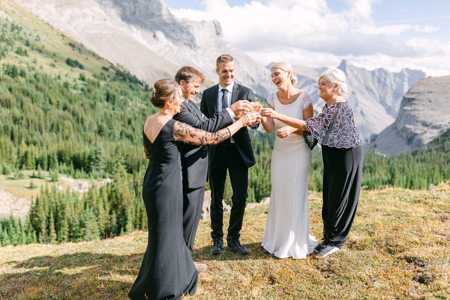 A joyful group of five people, including a bride in a white gown, raising glasses in a toast amidst a stunning mountain landscape.