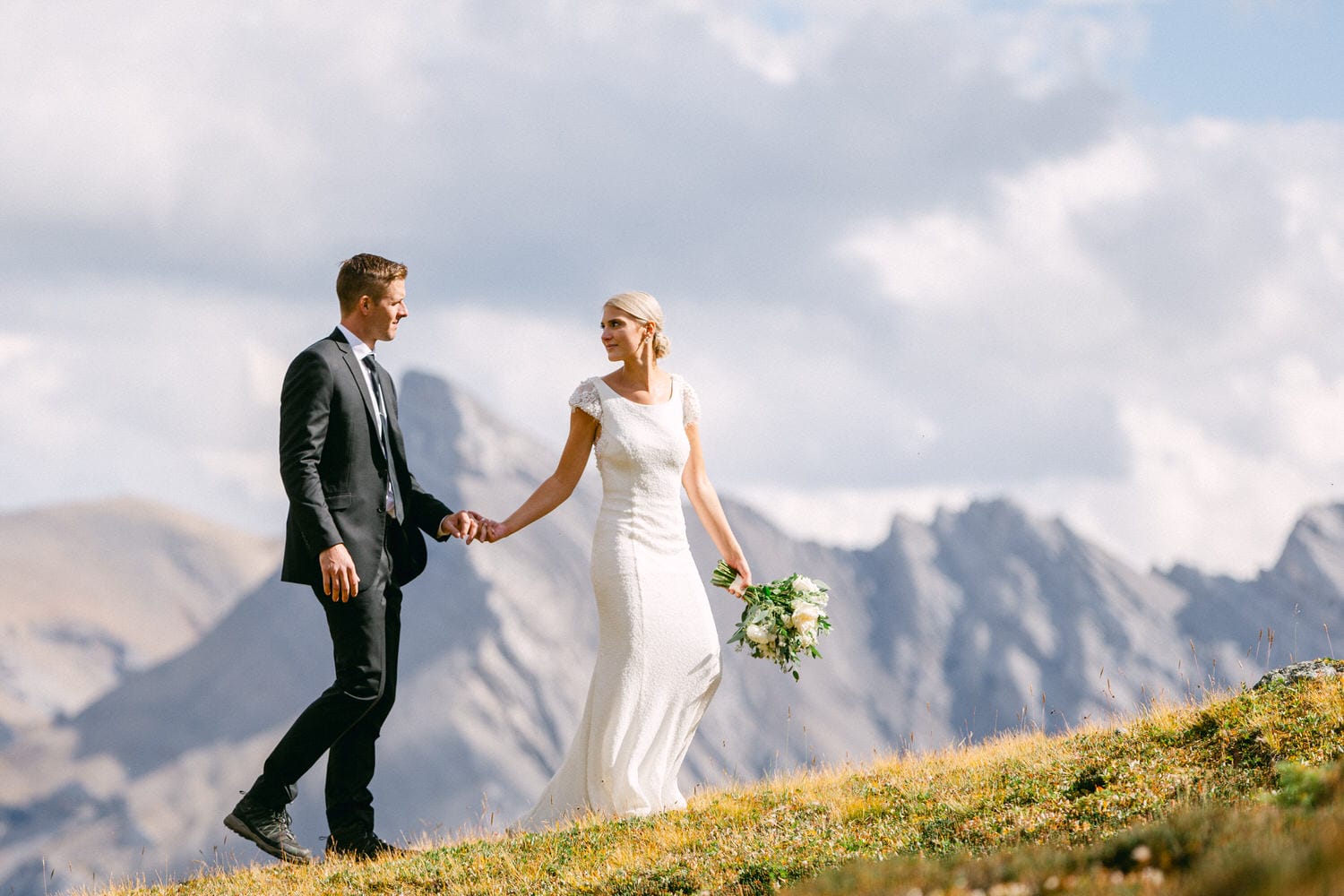 A bride and groom walking hand-in-hand on a grassy hillside with mountains in the background, capturing a moment of joy and romance.