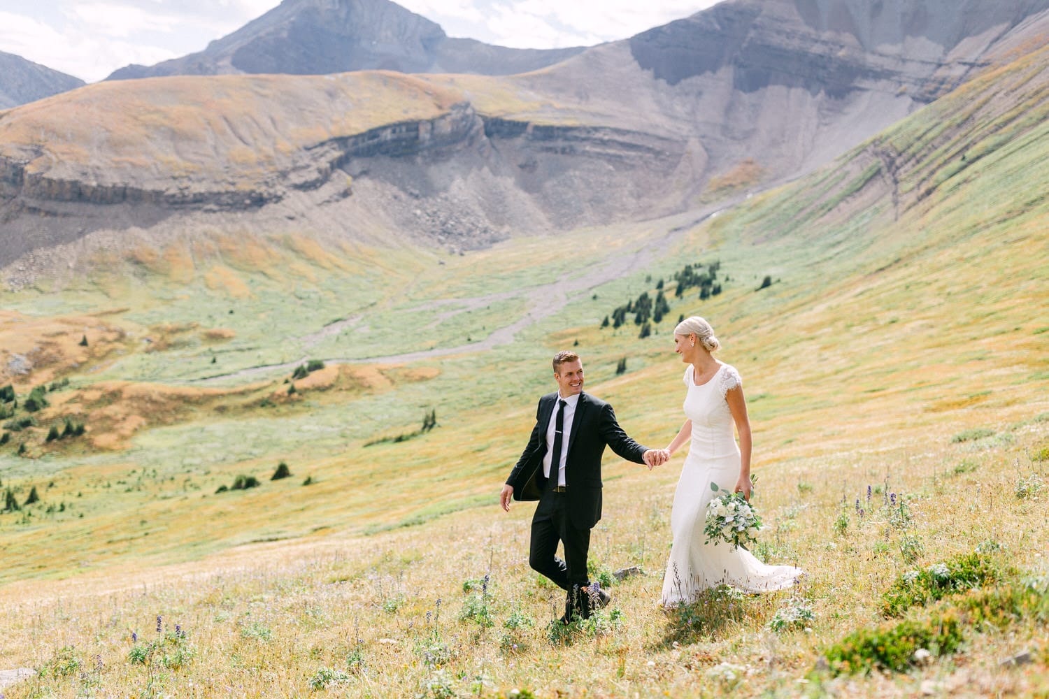 A happy couple walks hand-in-hand through a vibrant grassy landscape adorned with wildflowers, framed by majestic mountains in the background.