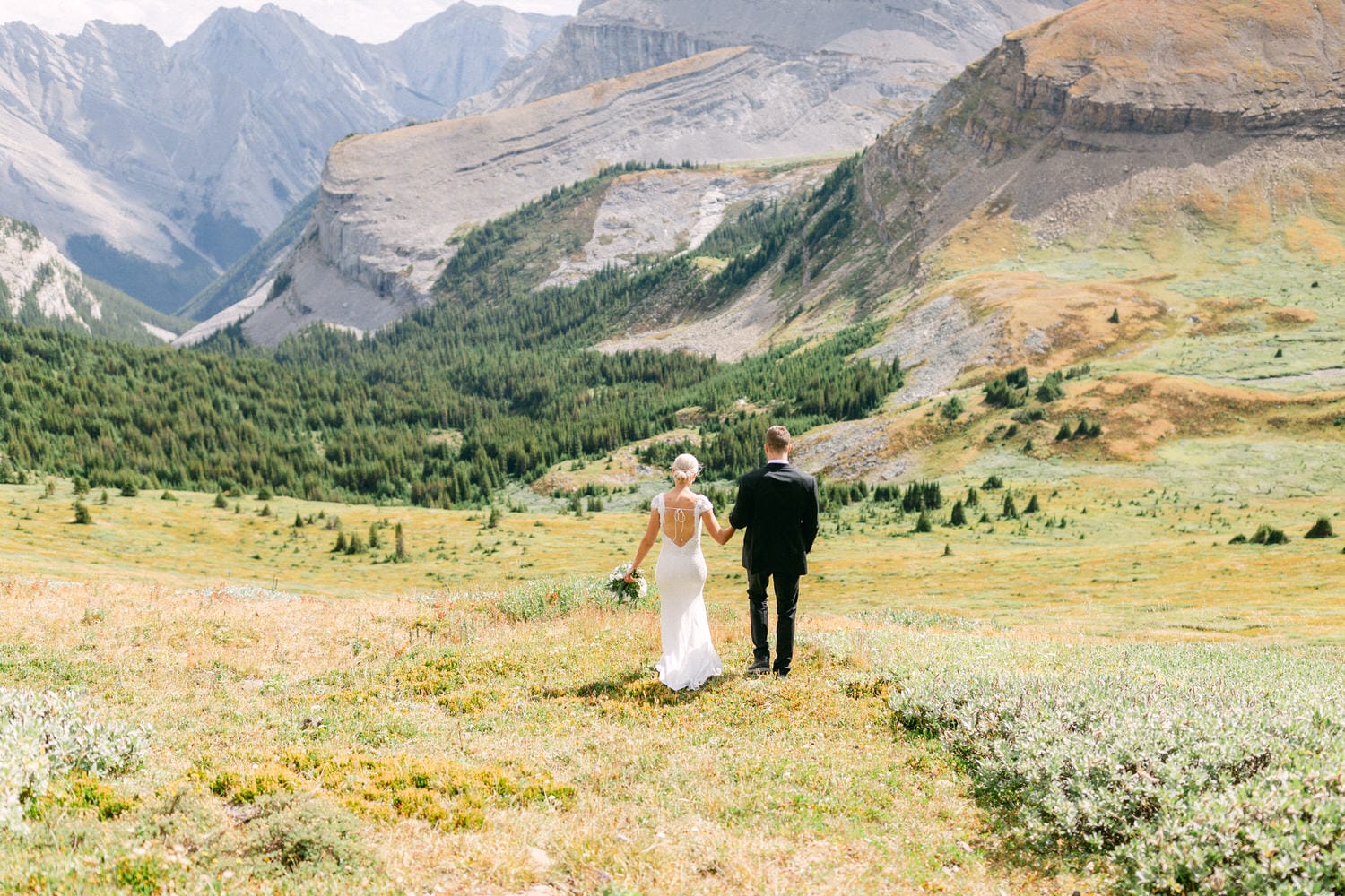 A bride and groom walking hand in hand through a scenic mountainous landscape, surrounded by greenery and golden fields.