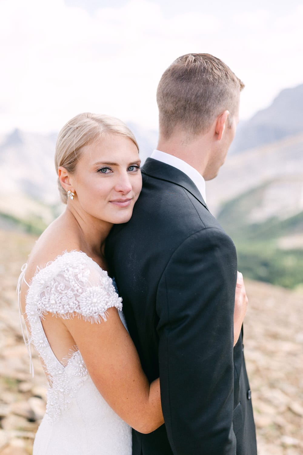 A bride gently embraces her groom from behind, showcasing her elegant wedding dress against a scenic mountainous backdrop.