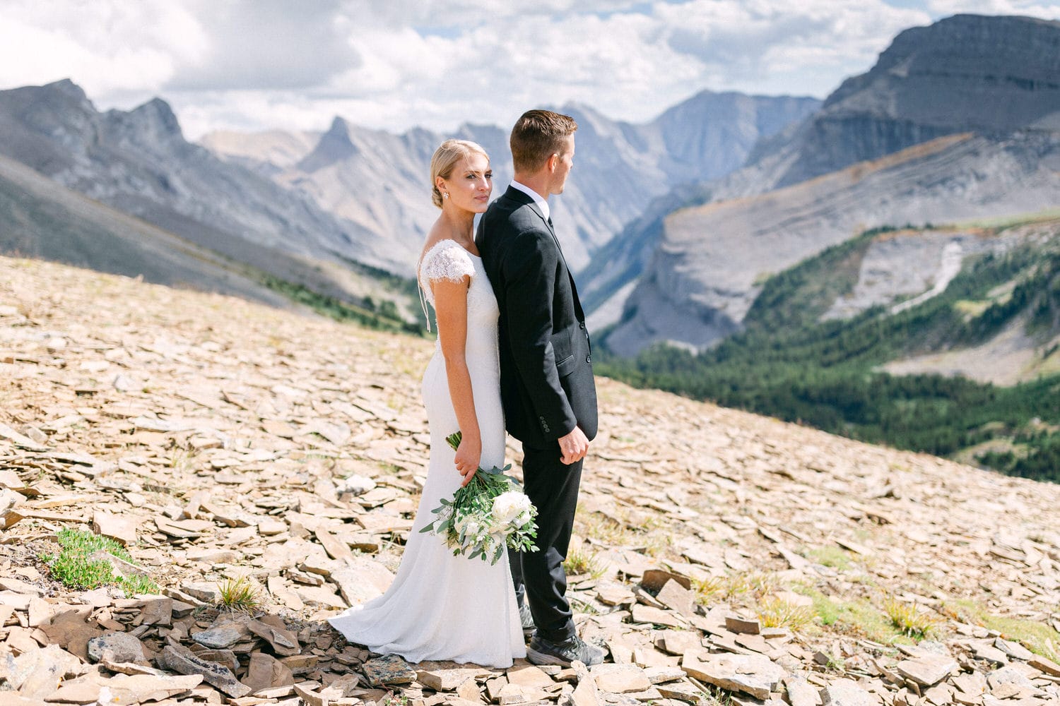 A bride and groom stand back-to-back in a stunning mountainous landscape, showcasing their love amid nature's beauty.