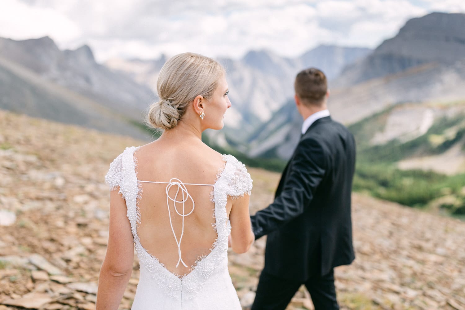 A bride with a backless dress and her partner are walking hand-in-hand amidst a stunning mountainous landscape.