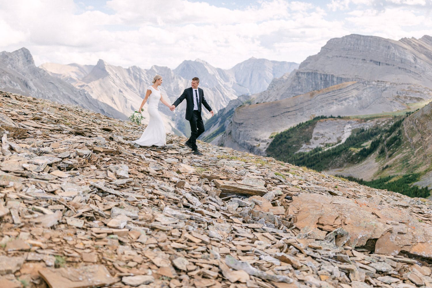 A couple joyfully walks hand in hand across rocky terrain, surrounded by stunning mountain peaks and a picturesque sky.