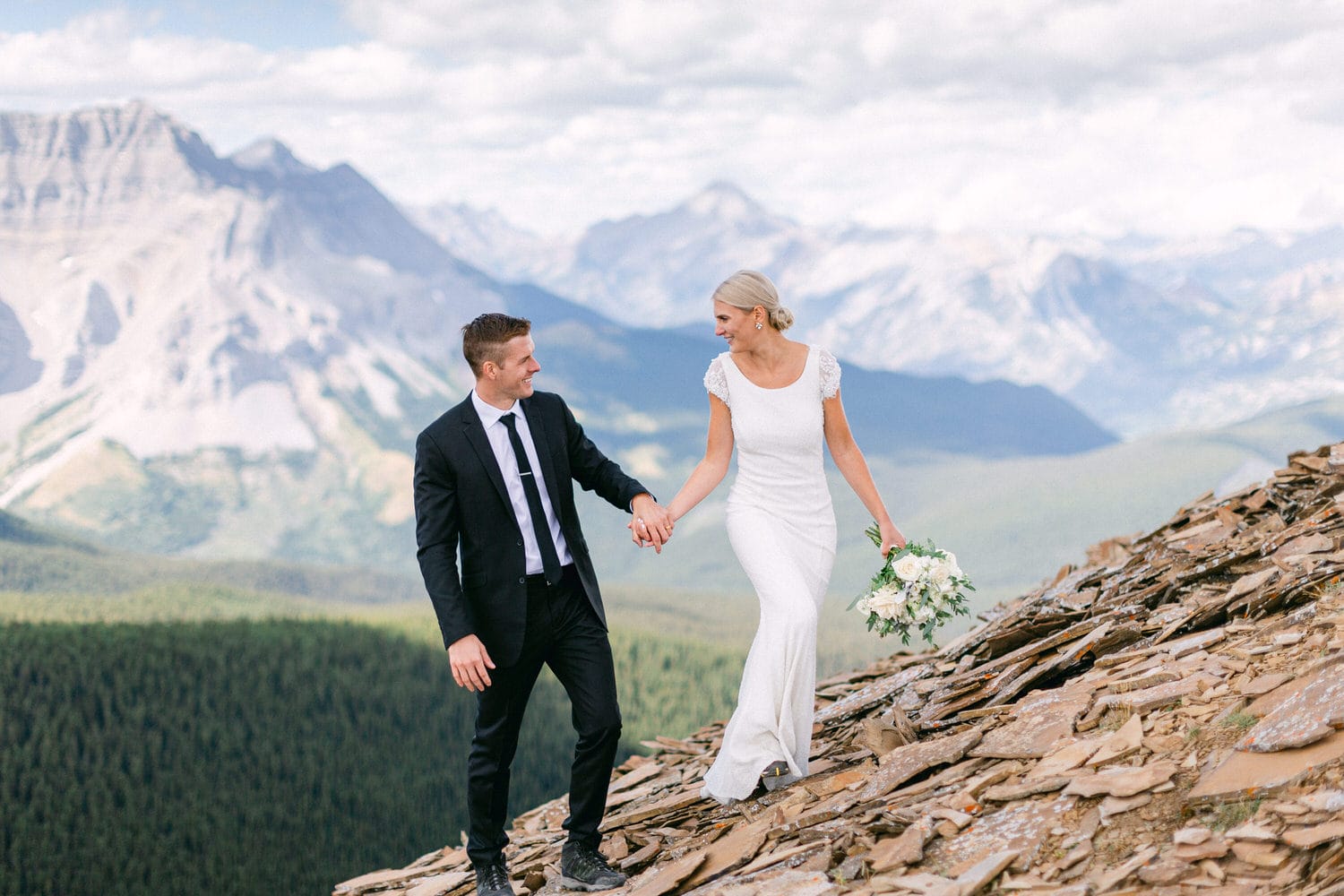 A couple joyfully walks hand-in-hand during their wedding ceremony atop a scenic mountain ridge, surrounded by lush landscapes and towering peaks.