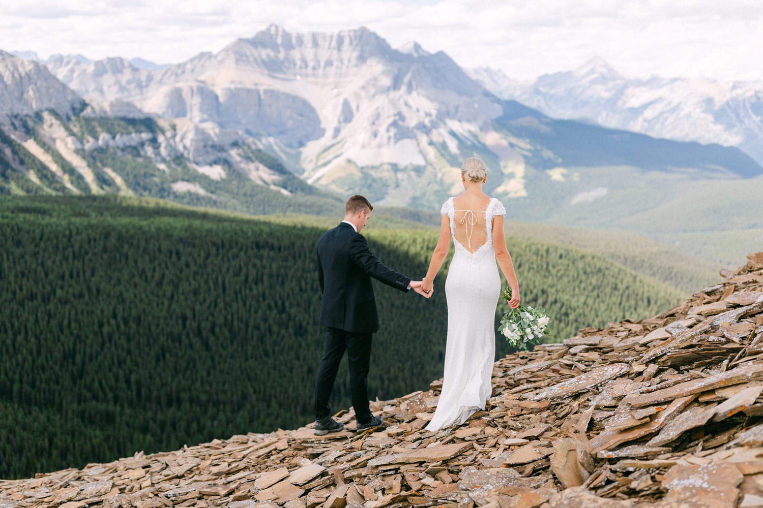 A bride in a flowing white dress and a groom in a black suit walk hand in hand along a rocky path with majestic mountains in the background.