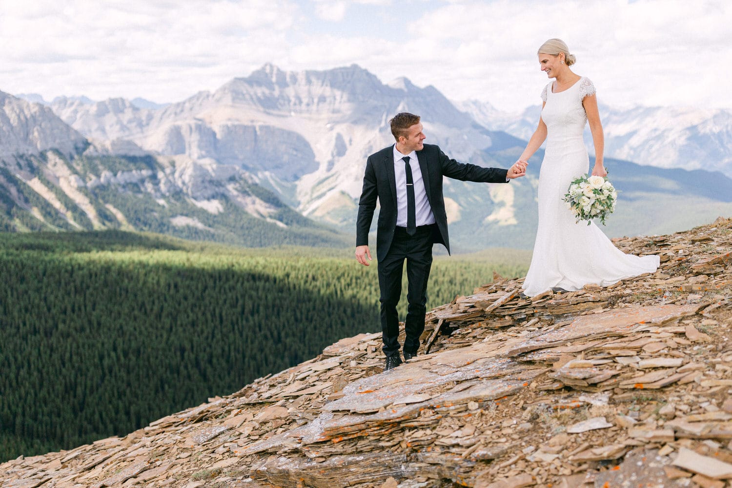 A bride and groom hold hands as they navigate rocky terrain on a mountain peak, surrounded by breathtaking landscapes.