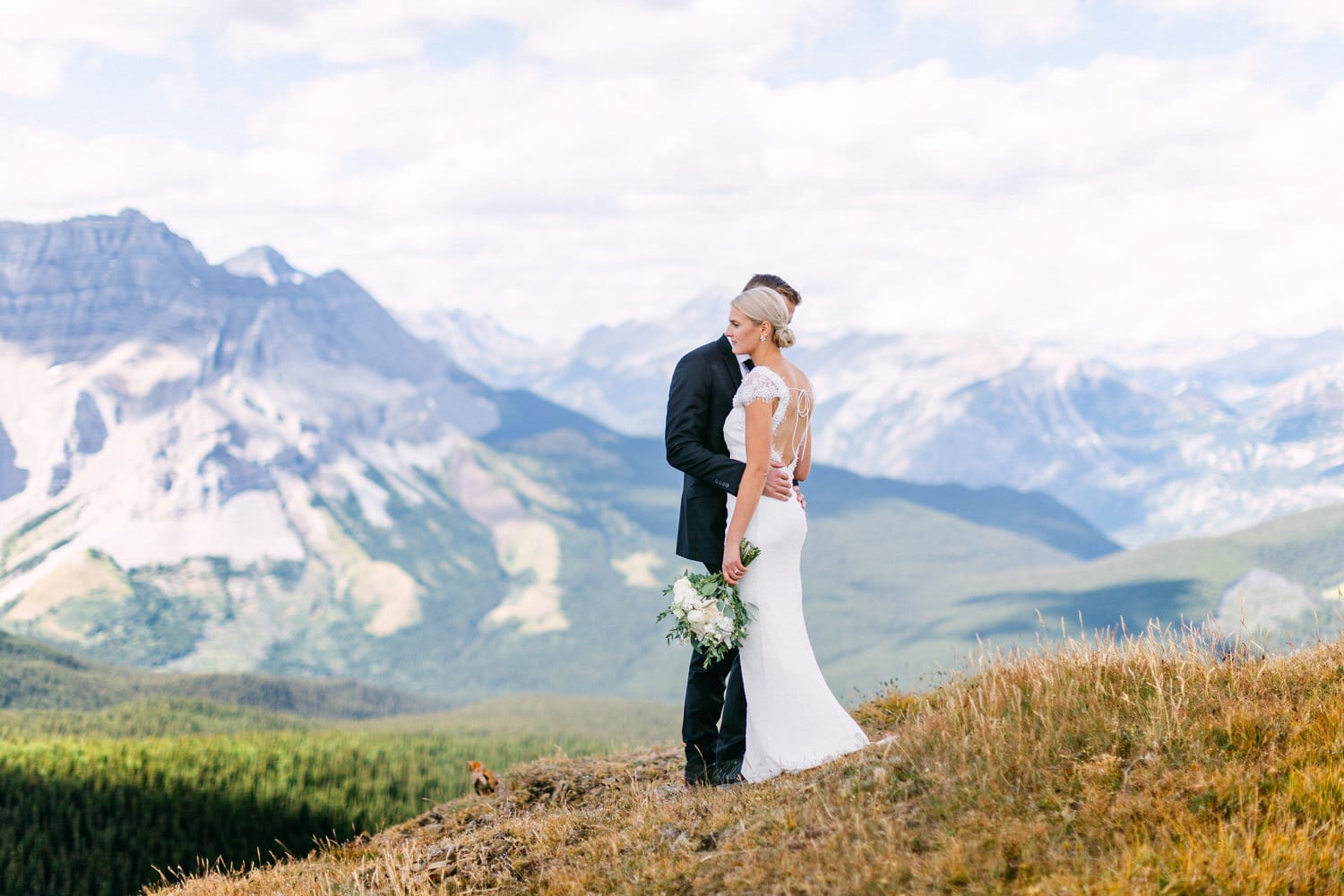 A bride and groom embrace on a mountain overlook, surrounded by breathtaking landscapes and vibrant greenery.