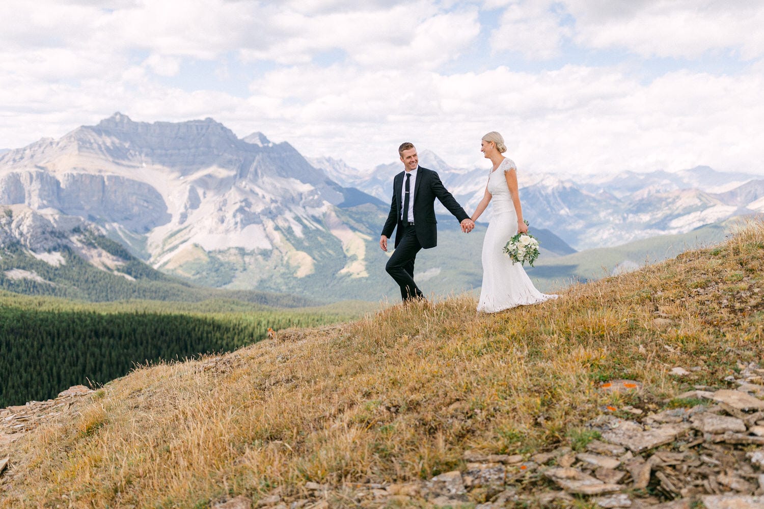 A bride and groom hold hands while walking on a mountain ridge, surrounded by stunning peaks and lush valleys.