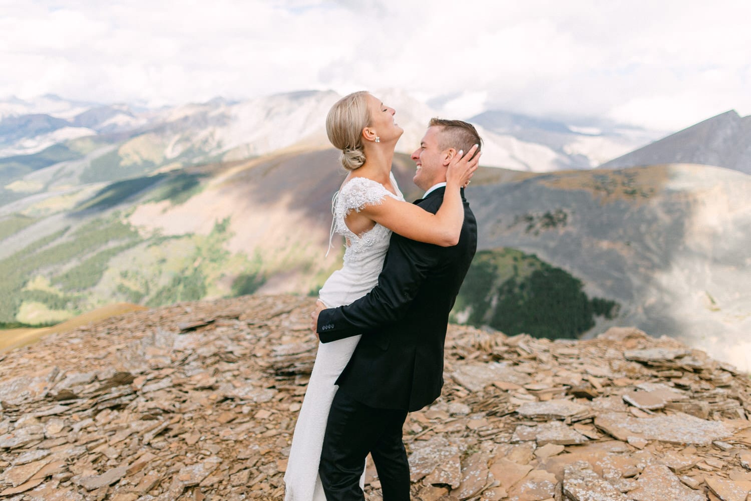 A joyful couple embraces on a rocky mountain peak, surrounded by stunning landscapes and dramatic skies.