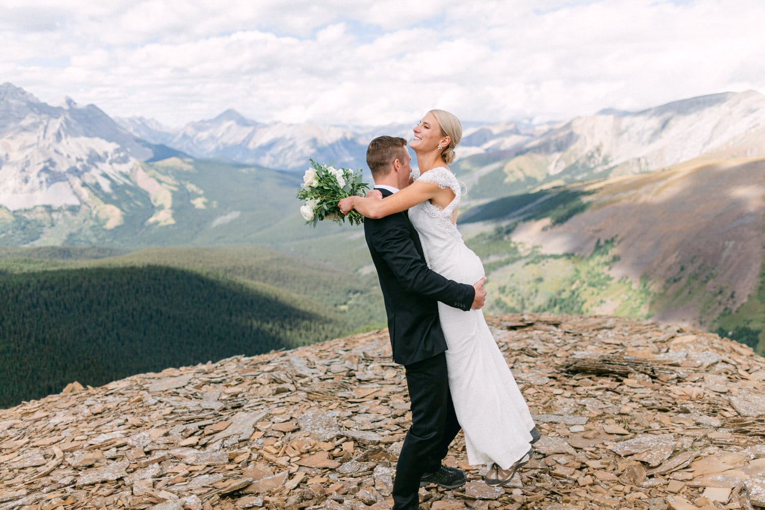 A groom lifts his bride in a stunning embrace atop a rocky mountain, surrounded by breathtaking landscapes and greenery.