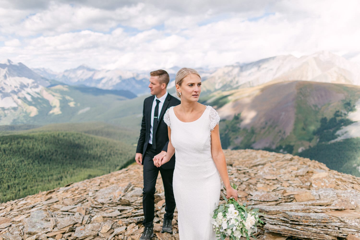 A bride and groom walking on a rocky ledge with a breathtaking mountain backdrop, dressed elegantly, surrounded by nature.