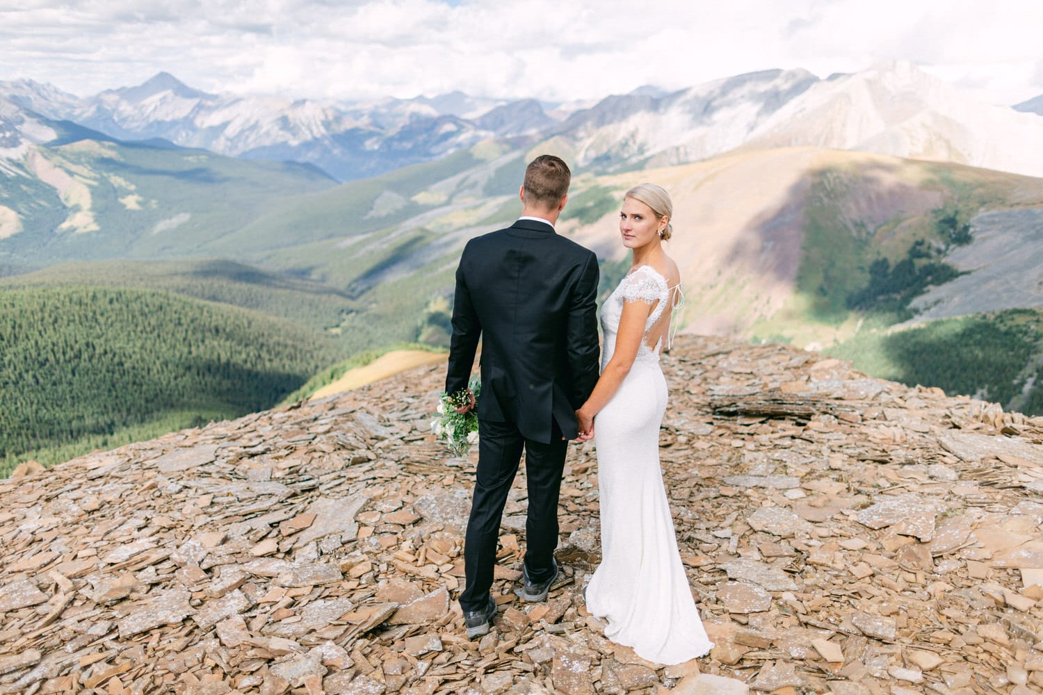 A couple dressed in formal wedding attire stands hand in hand on a rocky mountain overlook, surrounded by lush greenery and majestic peaks.