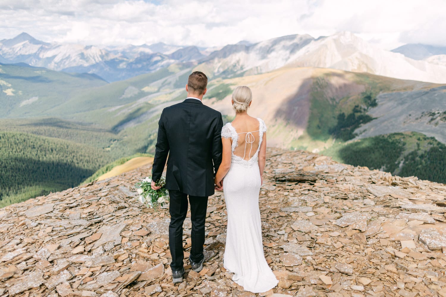 A couple stands hand in hand on a rocky cliff overlooking a breathtaking mountain landscape, embodying love and adventure.