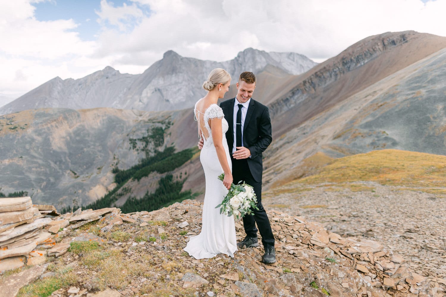 A couple stands on a rocky mountain top, exchanging joyful glances, surrounded by breathtaking mountainous scenery. The bride, in a fitted white gown, holds a bouquet, while the groom is dressed in a formal black suit.