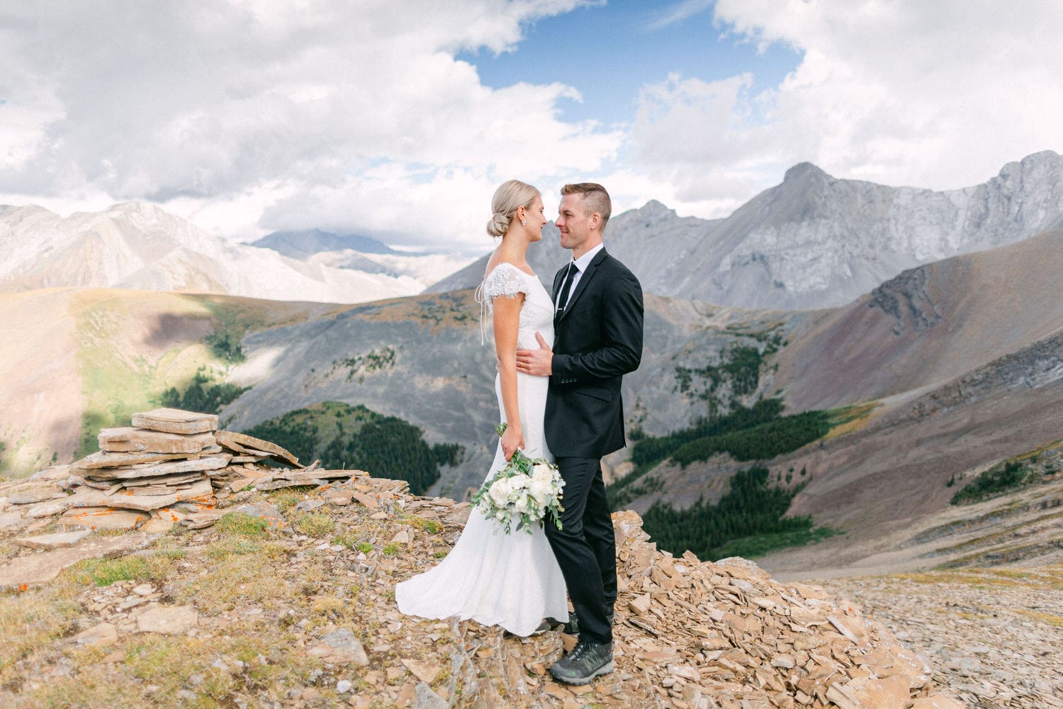 A bride and groom embrace on a rocky mountain peak, surrounded by stunning mountain vistas and dramatic clouds.