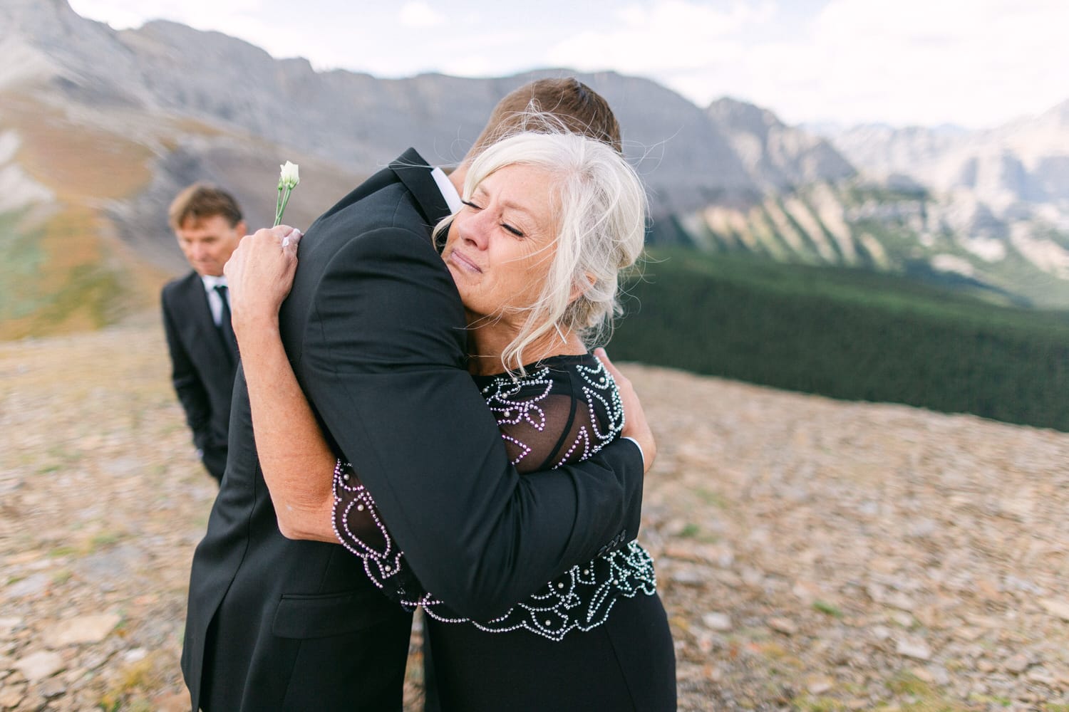 A heartfelt embrace between two individuals on a mountainside, with a backdrop of stunning peaks and greenery, capturing a moment of connection amidst a wedding celebration.