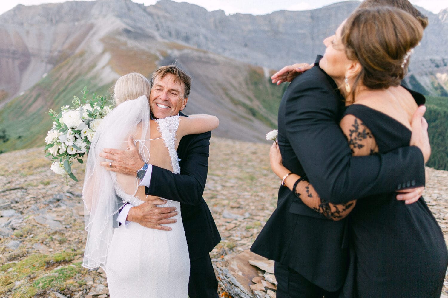 A joyful wedding scene featuring two couples hugging in a stunning mountain landscape, highlighting familial love and celebration.