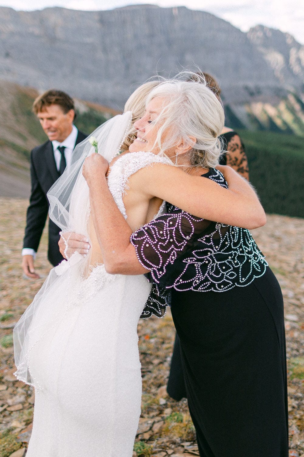 A bride in a white dress hugs a woman in a black beaded top against a scenic mountainous backdrop, capturing a heartfelt moment of celebration.