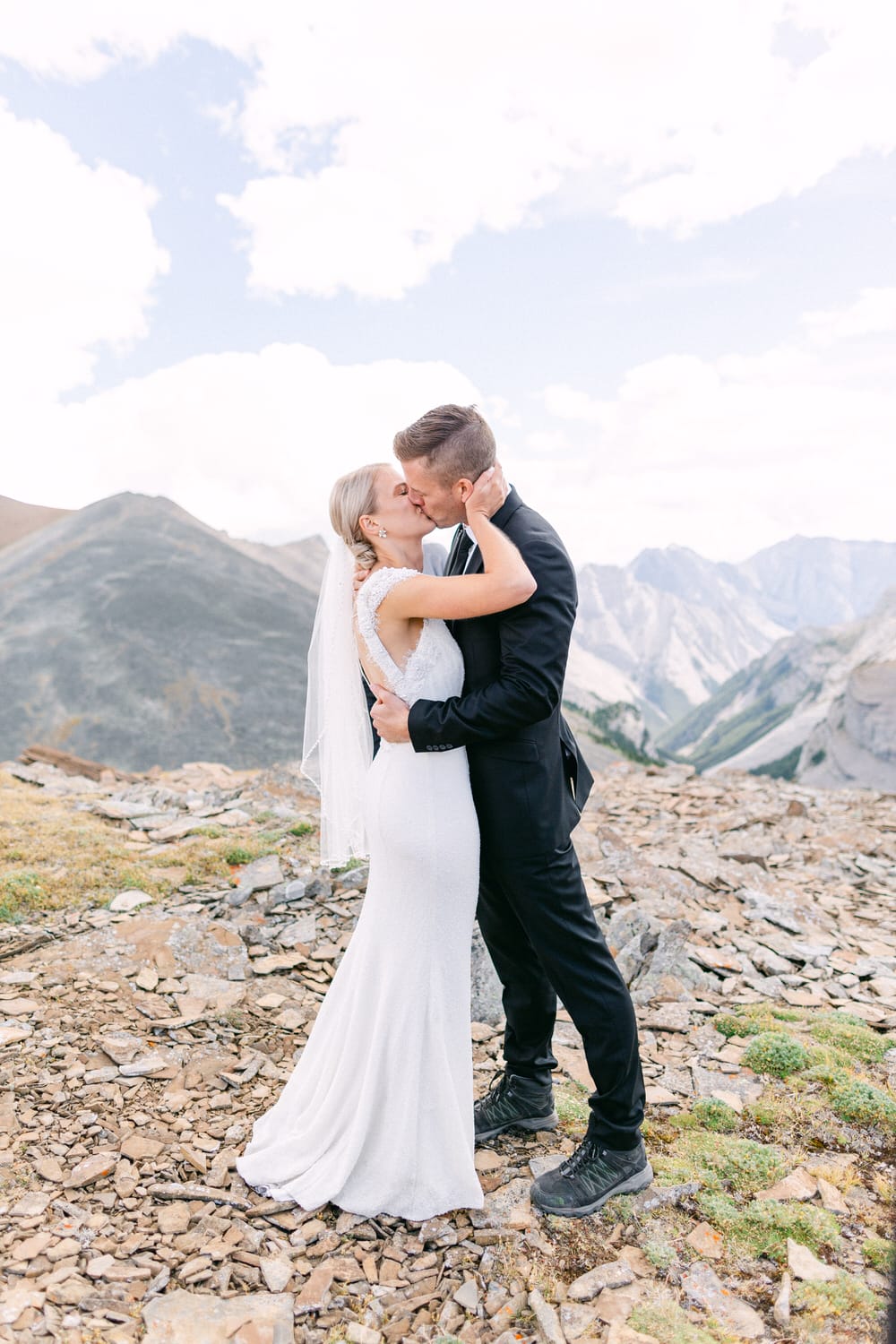 A couple sharing a kiss while standing on rocky terrain with mountains in the background, dressed in wedding attire.