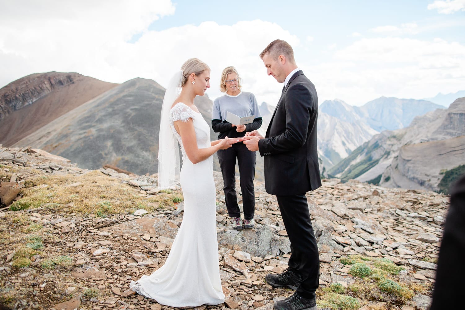 A couple exchanging vows on a mountain top, with a breathtaking view of the mountains in the background and an officiant present.