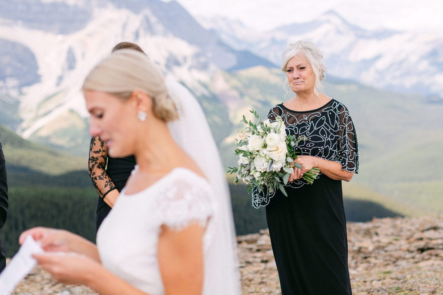 A bride reads her vows as her mother looks on, emotional, holding a bouquet amidst a stunning mountainous backdrop.