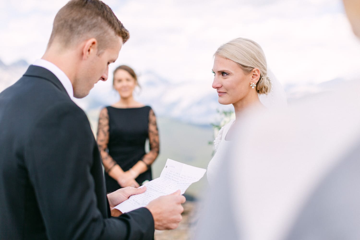 A couple exchanges heartfelt vows during an outdoor wedding ceremony, with the bride listening intently and a bridesmaid in the background.