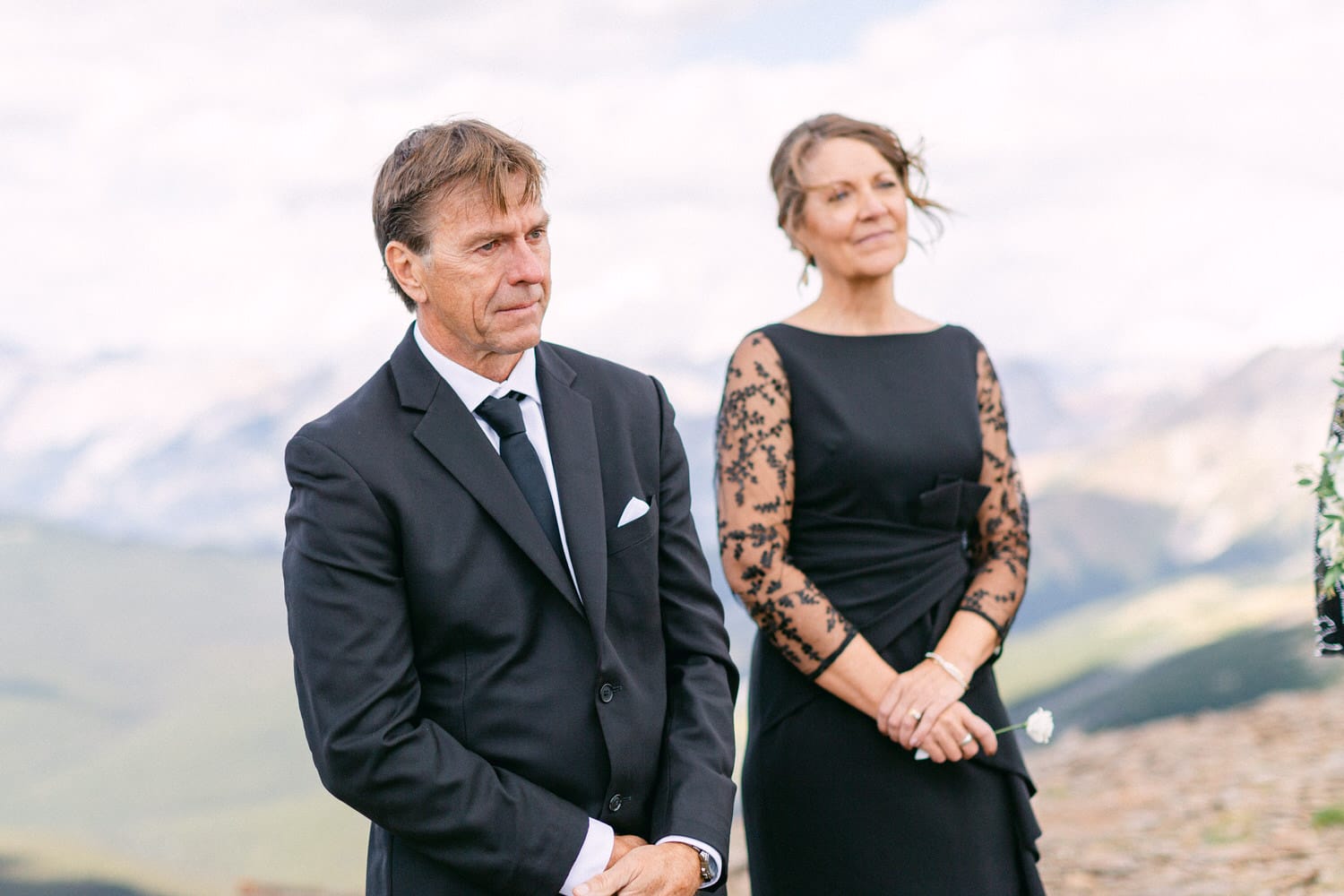 A couple stands together during a wedding ceremony in a scenic outdoor setting, with the bride's family members observing thoughtfully.