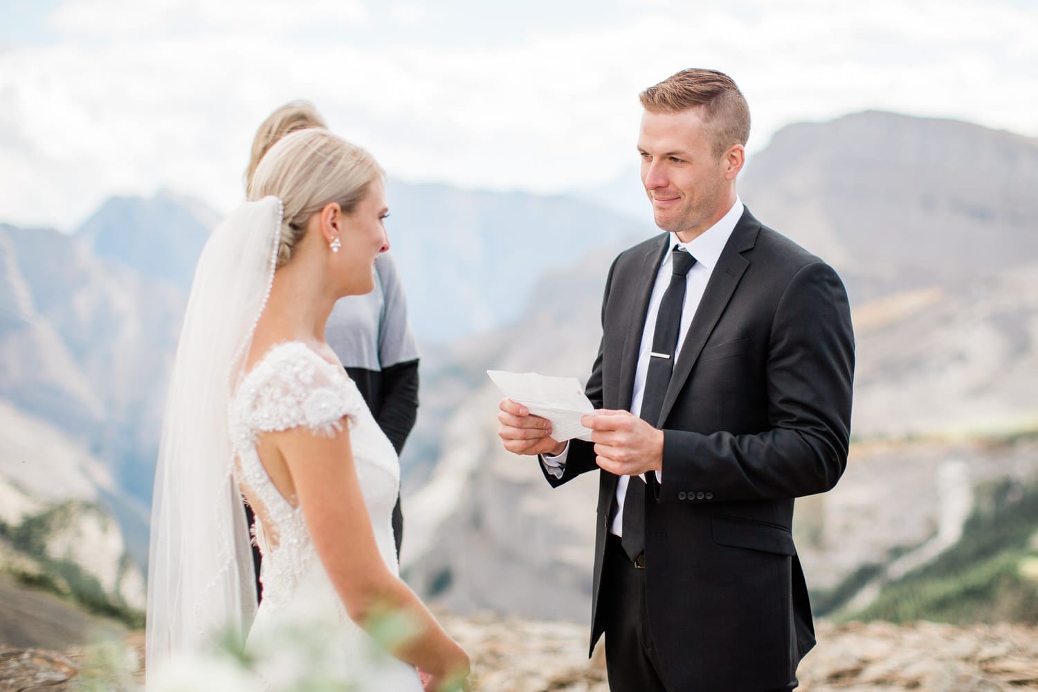 A groom reading vows to his bride during an outdoor wedding ceremony with a breathtaking mountain backdrop.