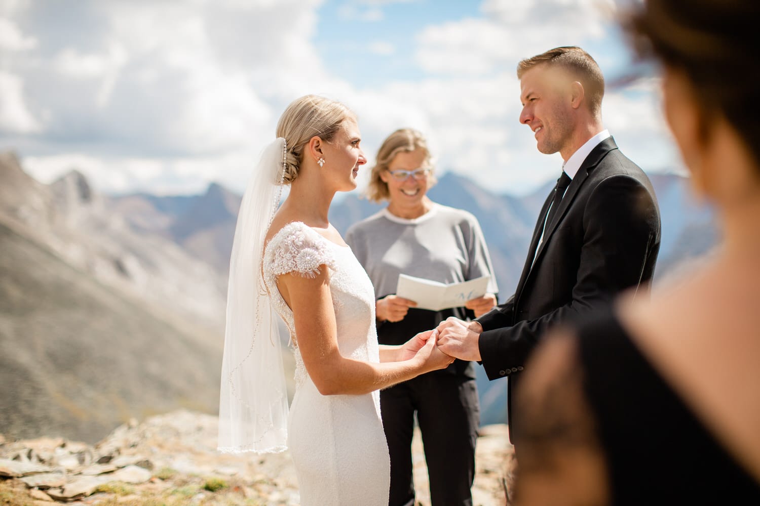 A bride and groom exchange vows during an outdoor wedding ceremony in a mountainous landscape, with a celebrant reading from a book.