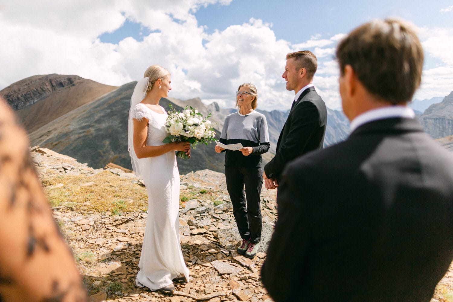 A bride in a white dress holds a bouquet while exchanging vows with her partner, surrounded by majestic mountains and a small group of guests.