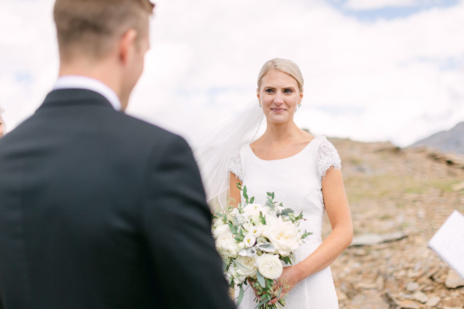 A bride with a bouquet smiles at her fiancé during an outdoor wedding ceremony set against a scenic landscape.