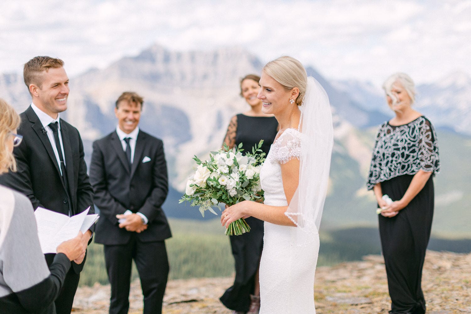 A bride smiles while holding her bouquet during an outdoor wedding ceremony in the mountains, surrounded by guests in formal attire.