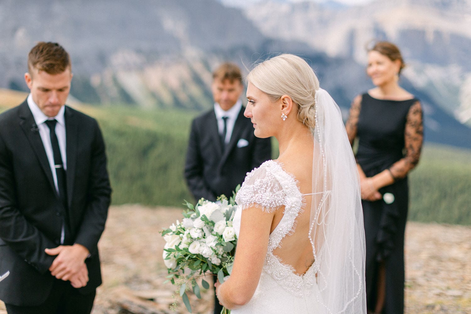 A bride stands with her back to the camera, holding a bouquet of white flowers, during a wedding ceremony in a scenic outdoor setting, accompanied by guests in formal attire.