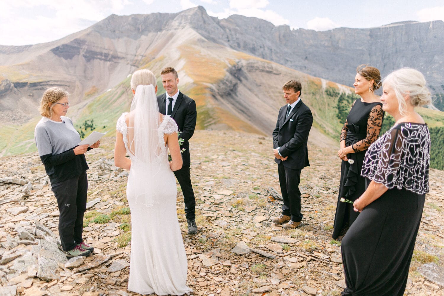 A bride and groom exchange vows in a scenic mountain setting, surrounded by guests and an officiant on a rocky terrain.