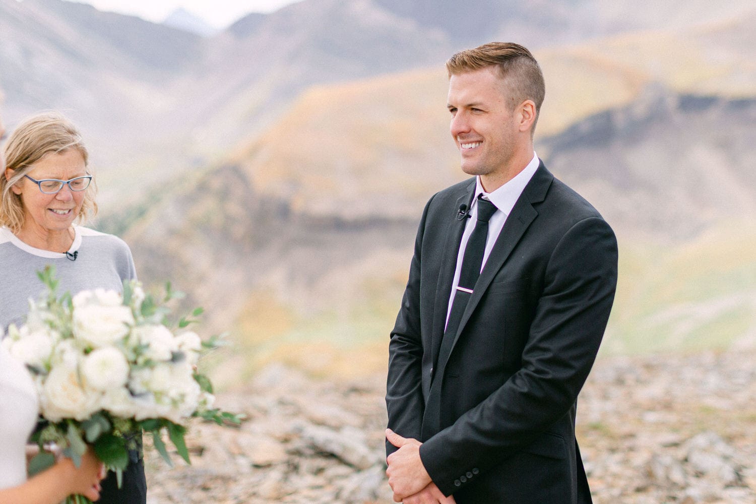 A man in a suit stands smiling during an outdoor wedding ceremony with a woman holding a bouquet and mountain scenery in the background.