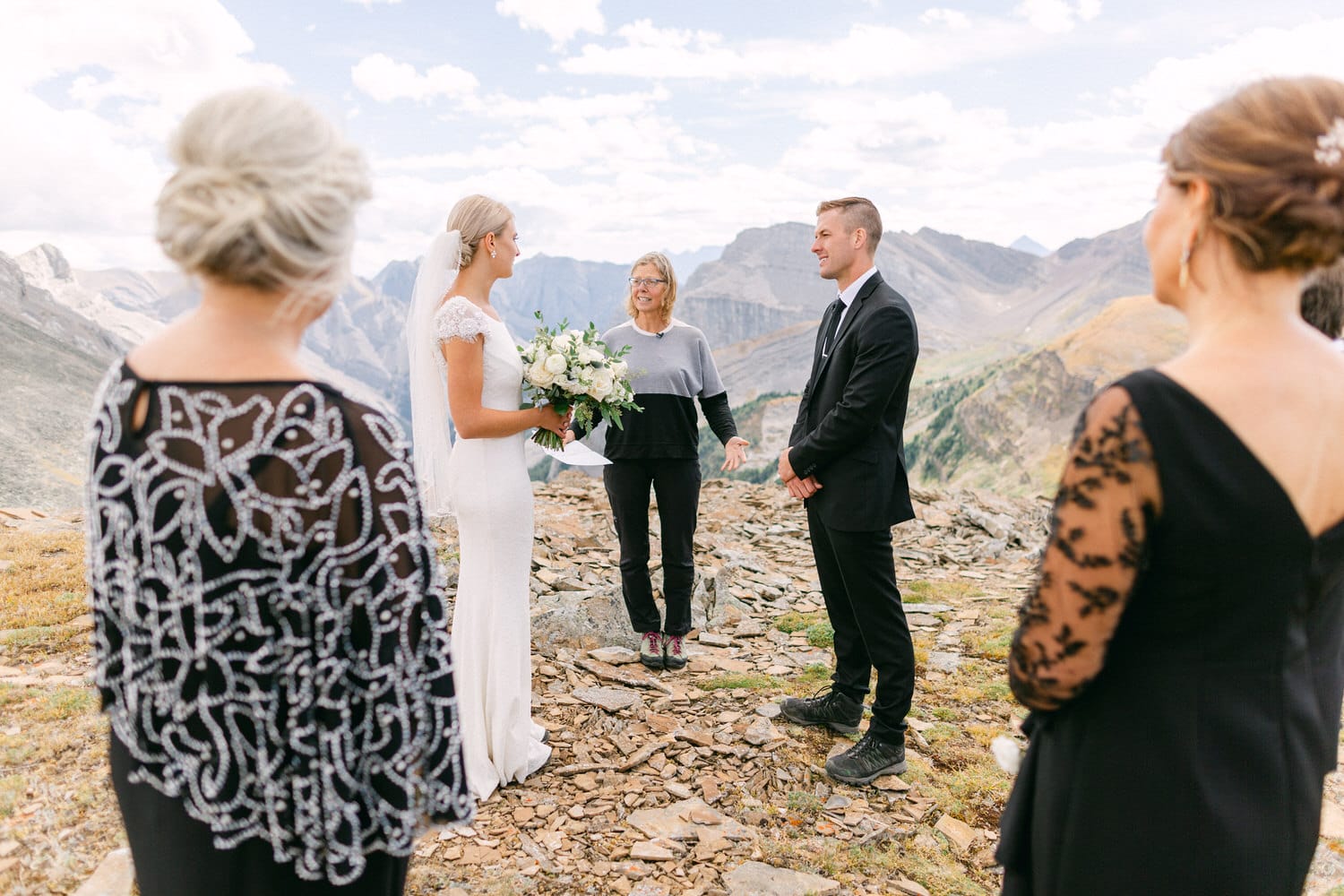 A bride and groom exchange vows at a scenic outdoor wedding surrounded by mountains, with guests observing in elegant attire.