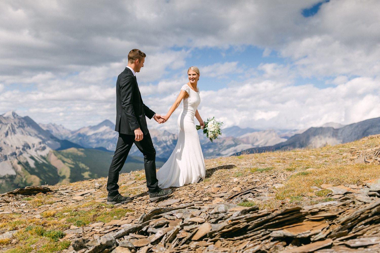 A couple walks hand in hand on a rocky mountain slope, surrounded by breathtaking mountain views, celebrating their wedding day.