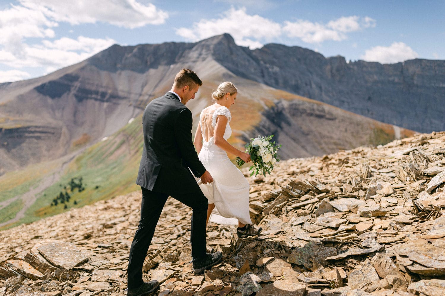 A couple dressed in wedding attire walks hand-in-hand over rocky terrain, with a stunning mountain backdrop under a bright sky.