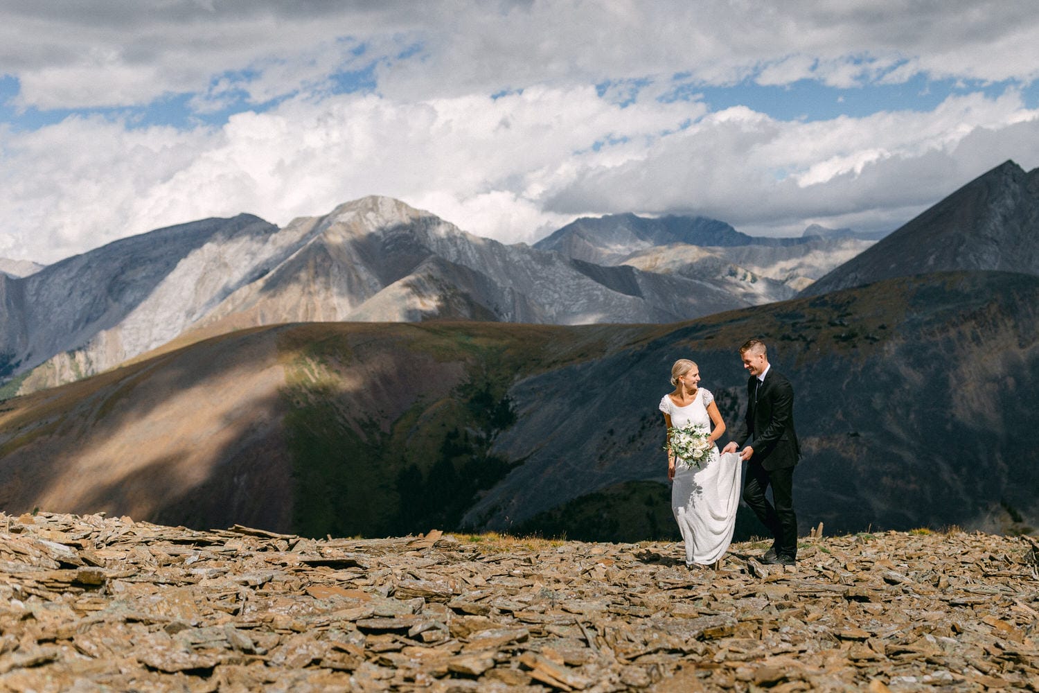 A bride and groom joyfully walk together on a rocky mountain landscape, surrounded by breathtaking peaks and a dramatic sky.