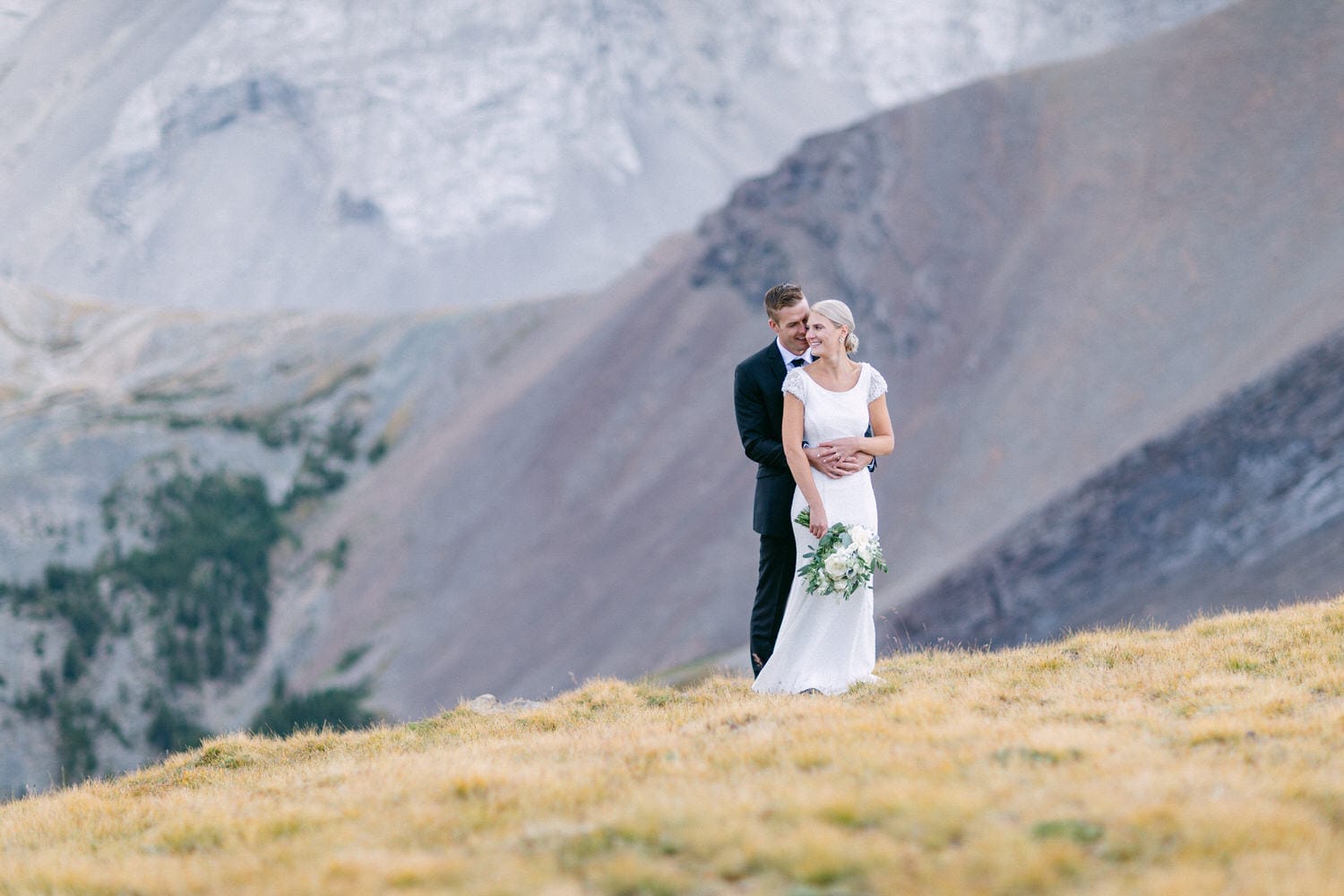 A bride and groom embrace on a grassy hillside with mountains in the background, showcasing their love in a serene outdoor setting.