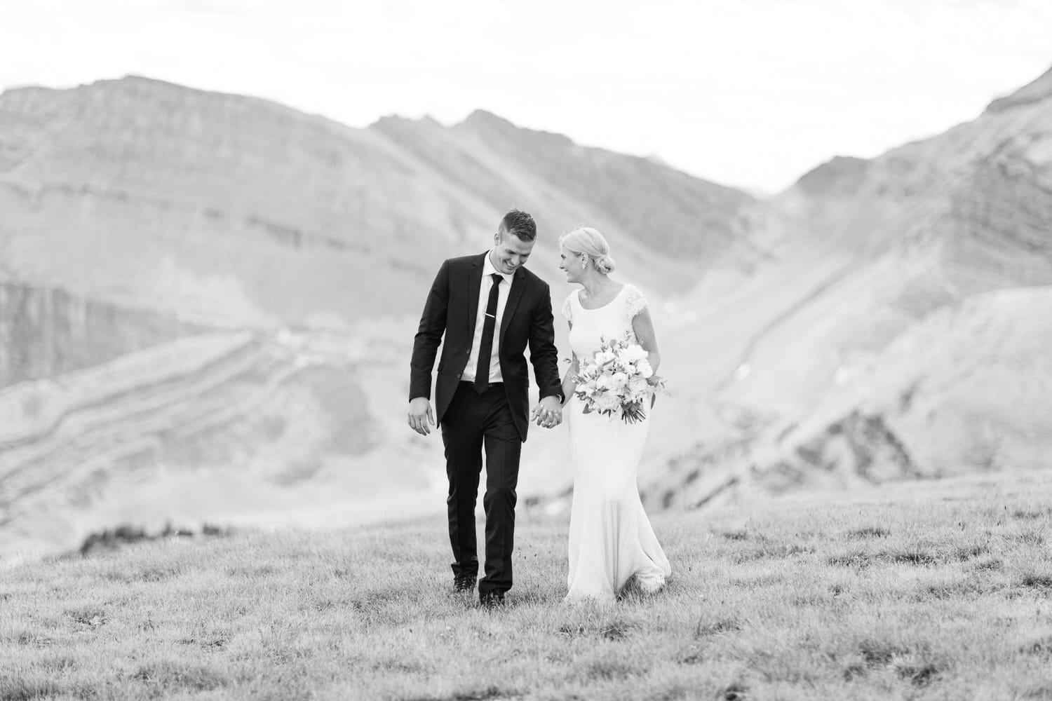 A happy couple walks hand in hand on a grassy hill with mountains in the background, capturing a romantic moment in black and white.