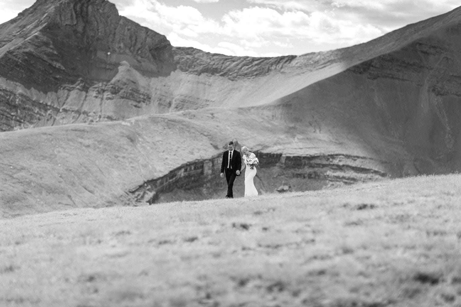 A couple walks hand in hand on a grassy hill with majestic mountains in the background, capturing a serene moment in their wedding journey.