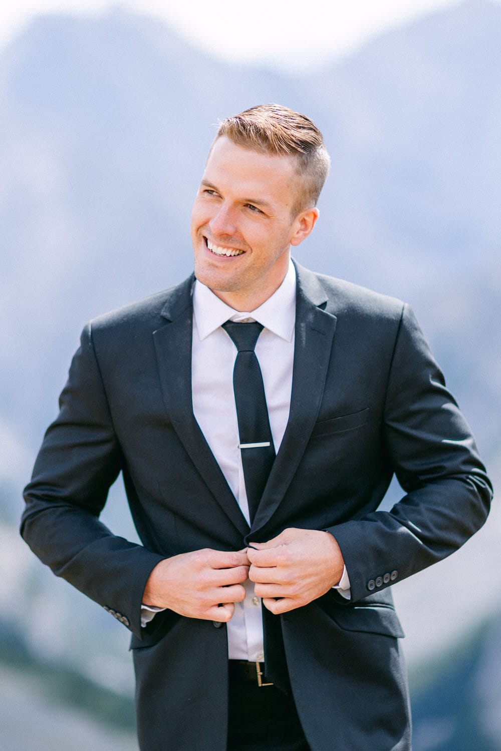 A cheerful man in a black suit and tie smiles while adjusting his jacket against a mountainous backdrop.