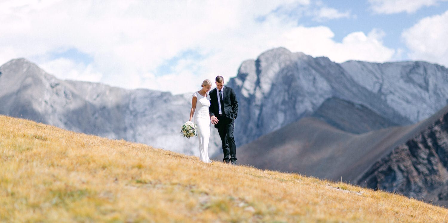 A bride and groom share a tender moment on a grassy slope, surrounded by stunning mountain scenery.