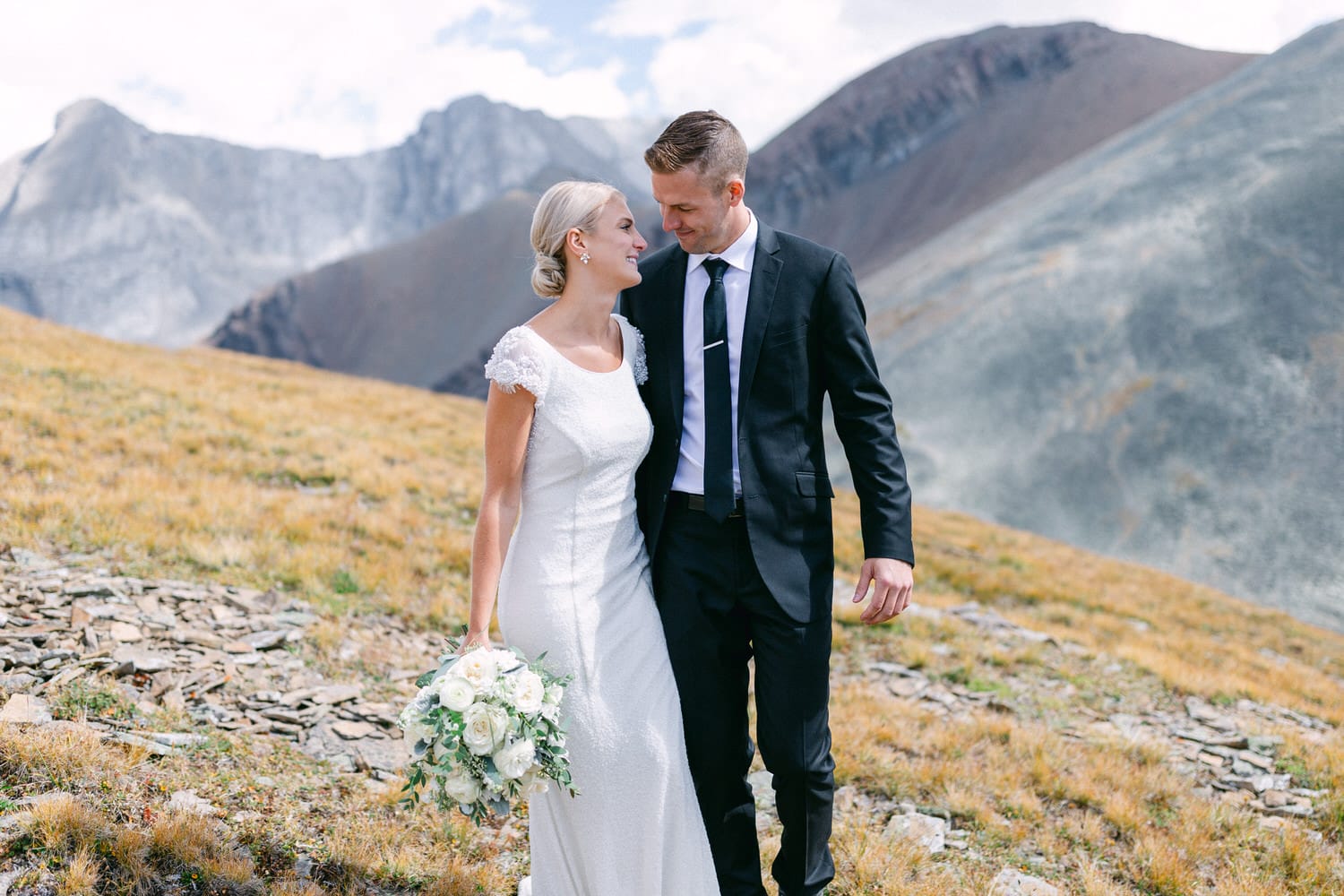 A happy couple in formal attire poses on a mountain landscape, with the bride holding a bouquet and the groom gazing at her warmly.