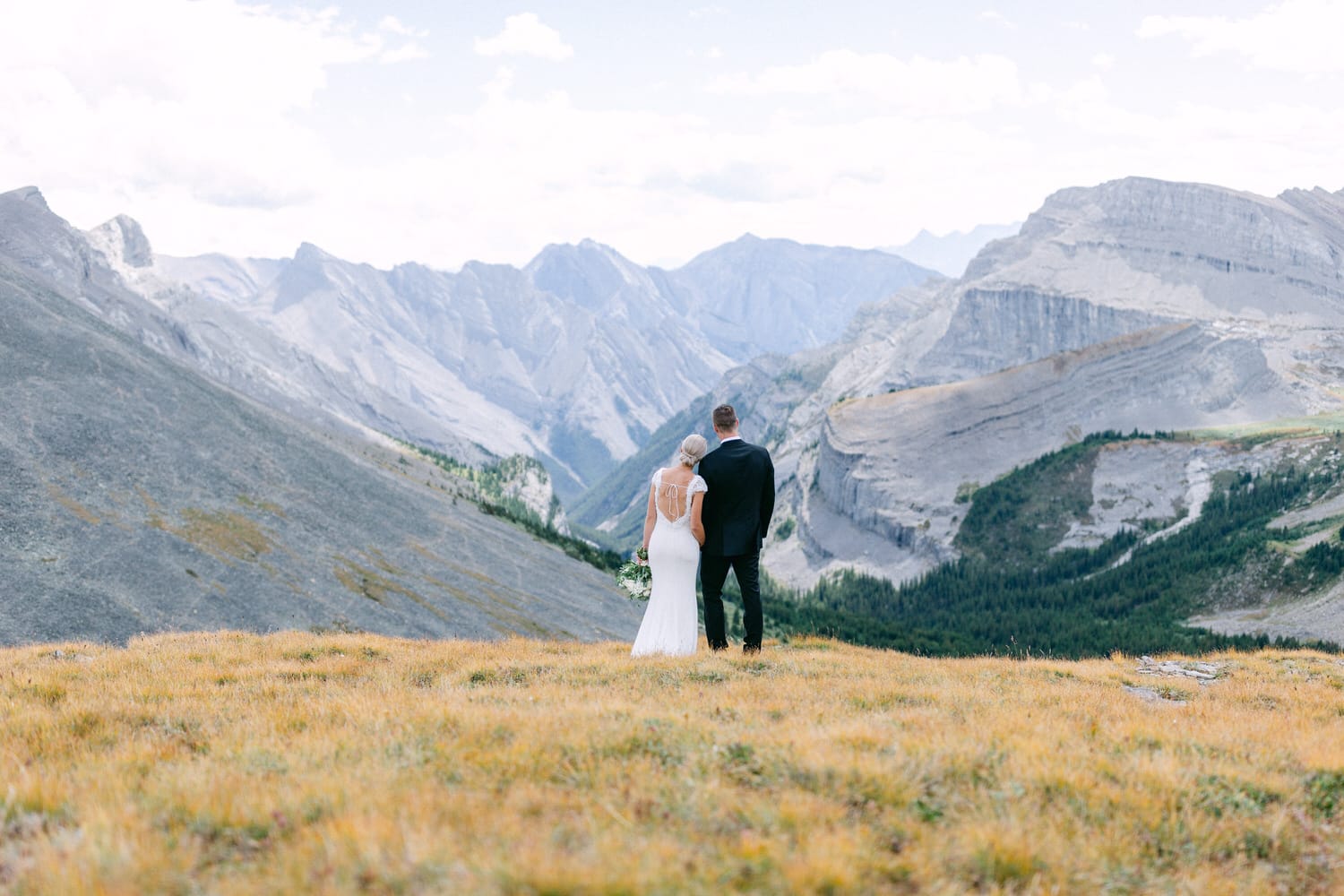 A couple stands hand in hand on a mountainside, overlooking dramatic peaks and valleys in a serene wedding moment.