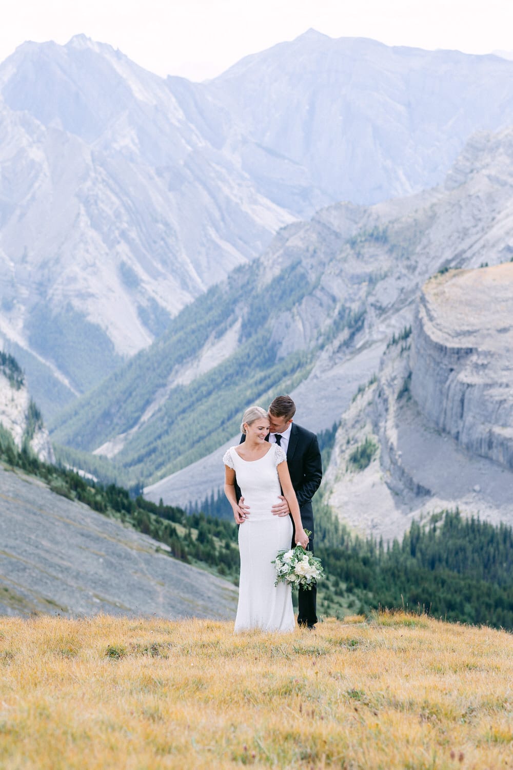 A couple embracing in a scenic mountain landscape, surrounded by lush greenery and rocky peaks, capturing a moment of love on their wedding day.