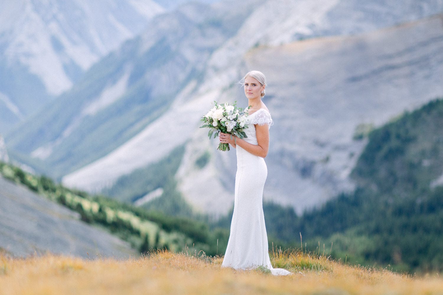 A bride stands gracefully on a grassy knoll in the mountains, holding a bouquet of white flowers against a stunning backdrop of rocky peaks and green valleys.