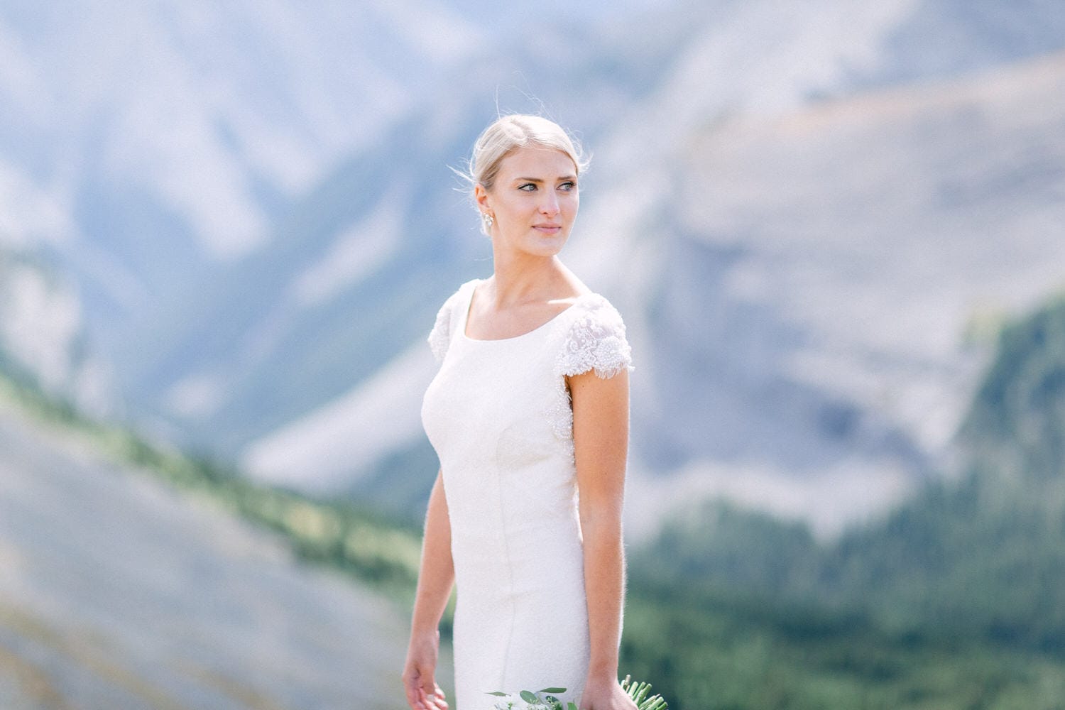 A bride wearing a white dress with lace sleeves stands gracefully against a backdrop of mountains, holding a bouquet in her hand.